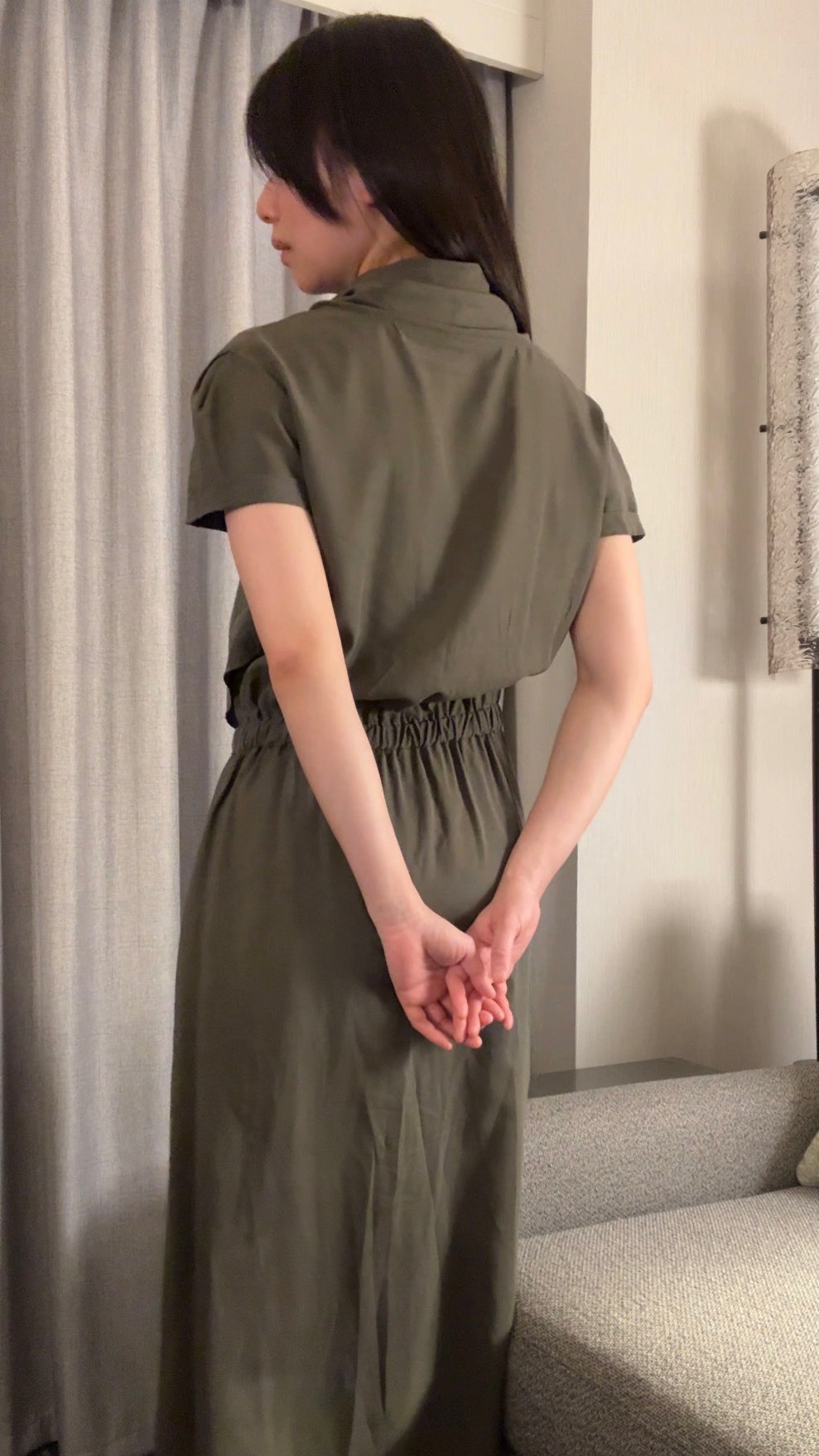 Woman wearing a green dress standing in a room with a gray curtain and sofa.