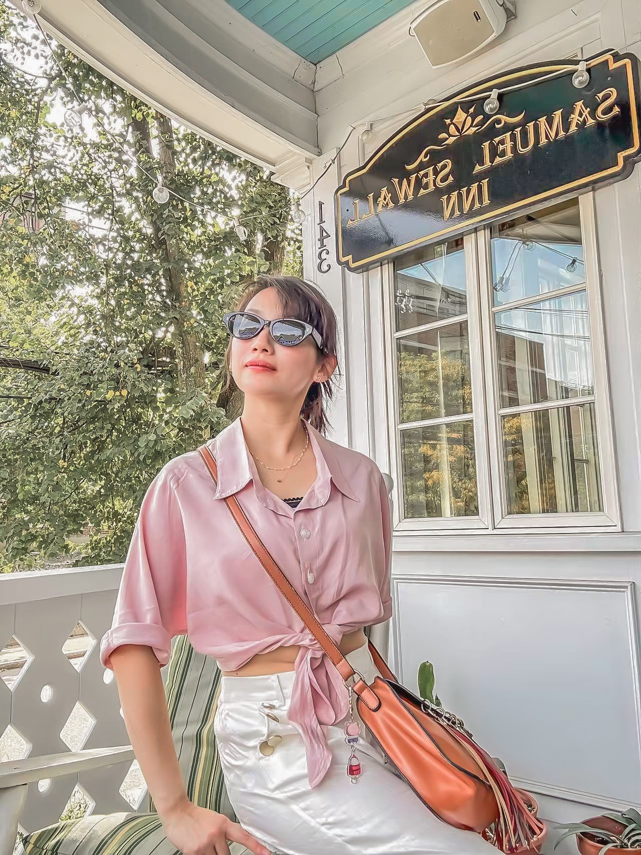 Woman in pink shirt and white pants standing on a porch with 'Samuel Sew & Inn' sign in the background