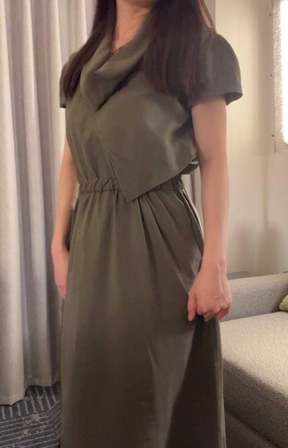 Person wearing a green dress standing in a room with curtains and a sofa.